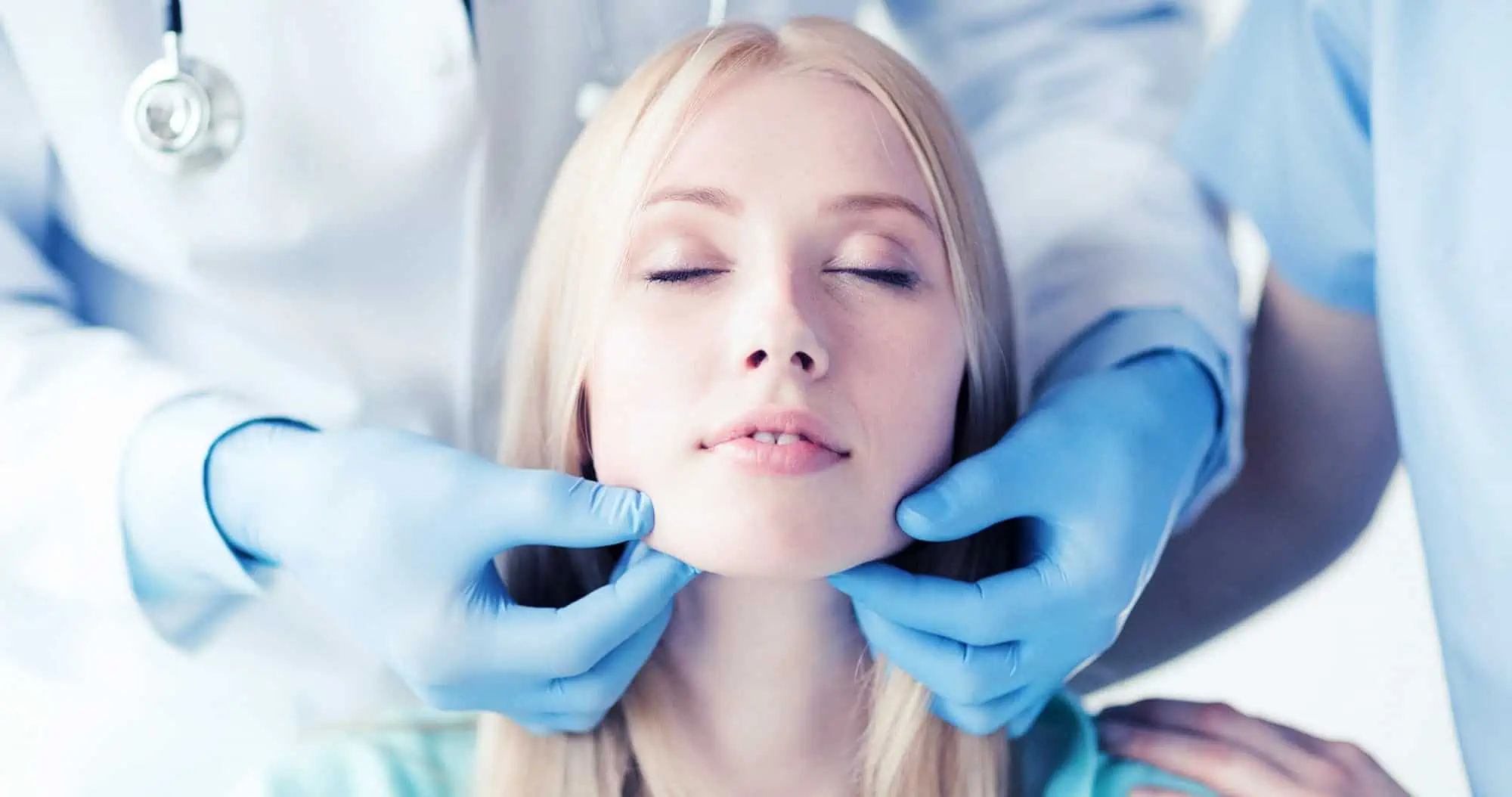 A young female patient's jawline is assessed by a skilled doctor wearing blue gloves.
